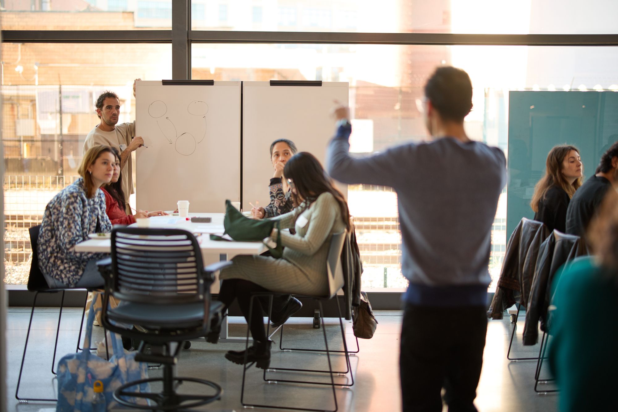The workshop presenters speak to the group, one of them drawing on the whiteboard. Tables look on attentively.